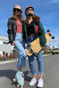 Zwei Frauen mit Skateboard und Rollerskatern stehen unter einem blauen Himmel