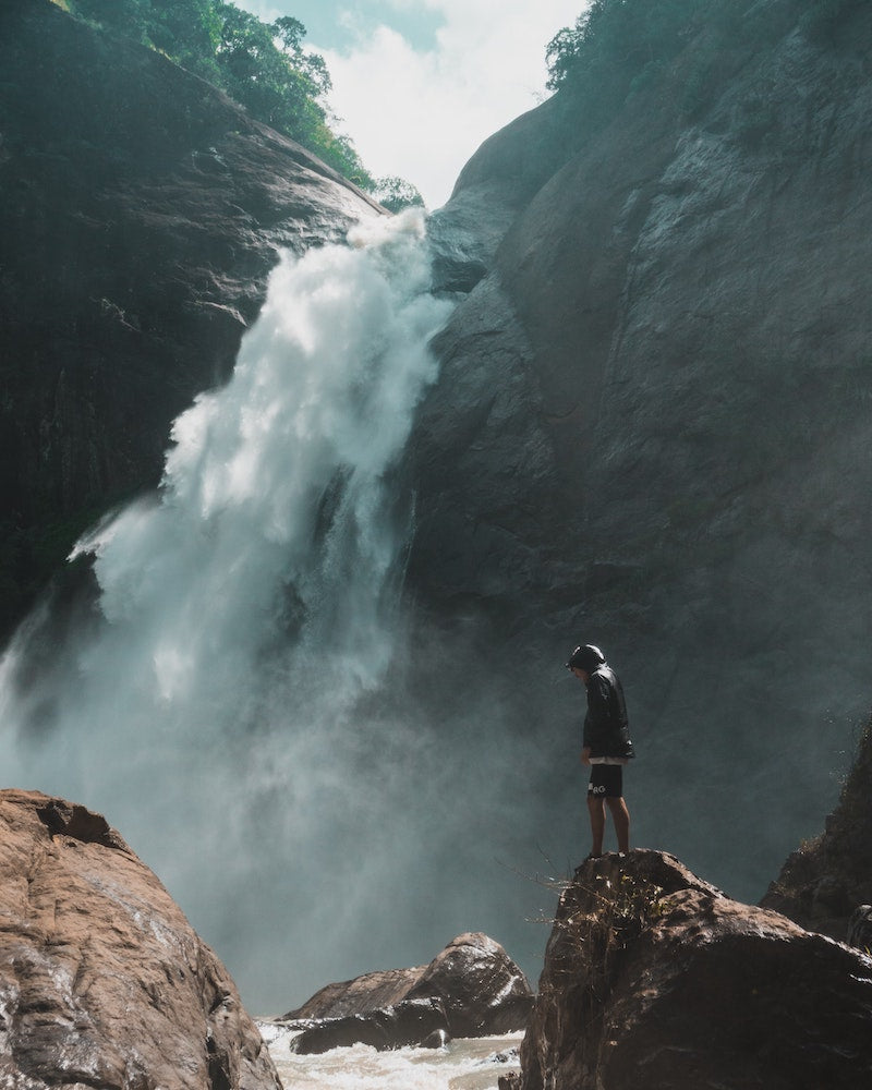 Im Hintergrund ist ein Wasserfall. Im Vordergrund steht rechts ein Mann auf einem Felsen. 