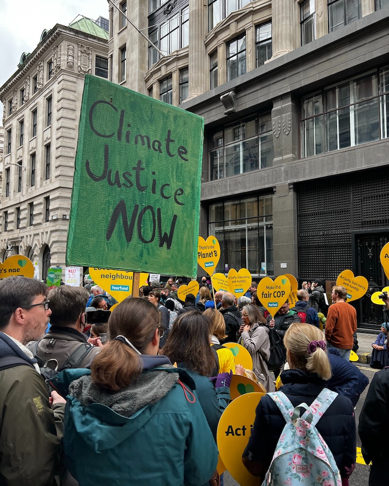 Mittig ist ein "Climate Justice Now" Schild in Grün. Im Vordergrund stehen Menschen bei einer Demonstration. 