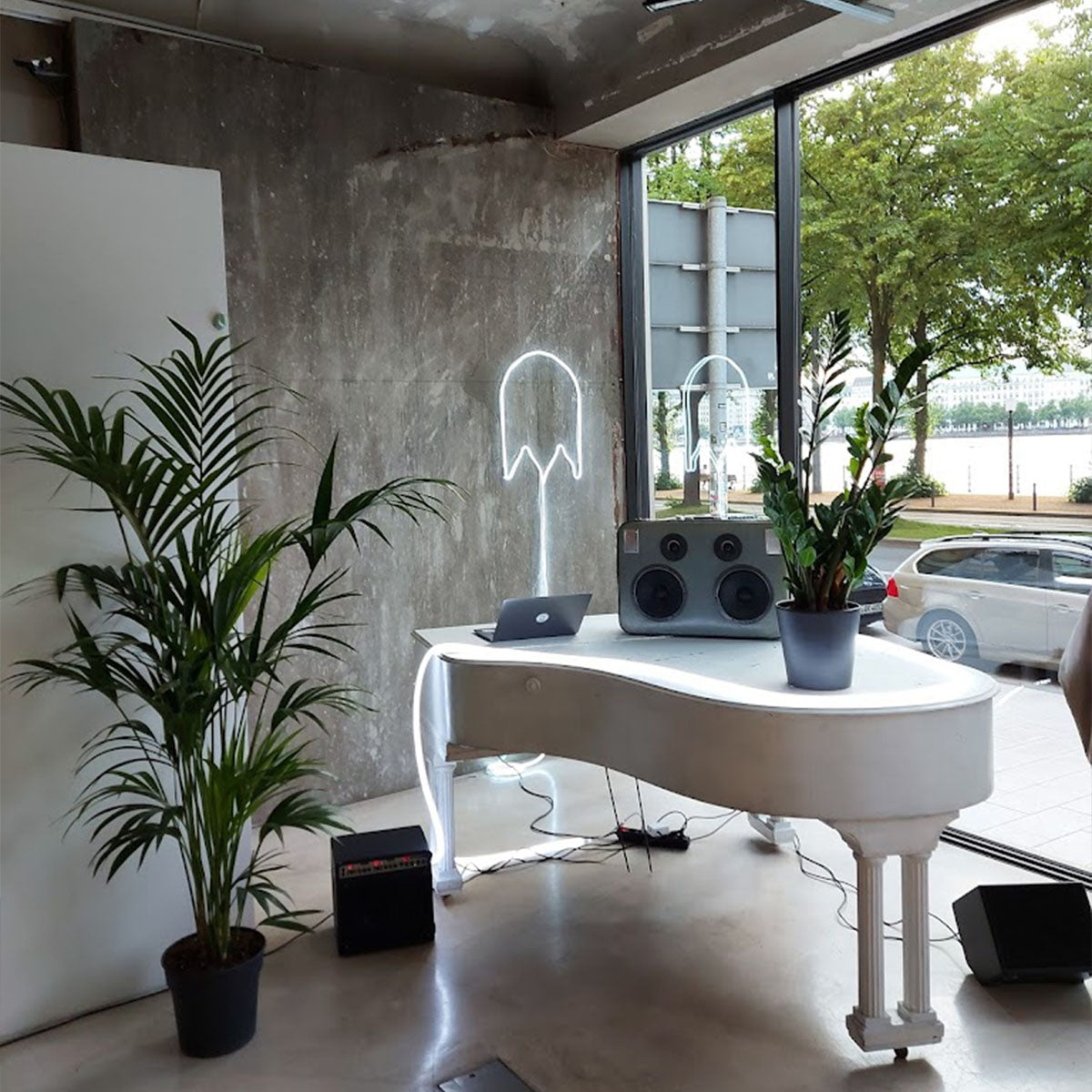 Links steht eine Topfpflanze. Rechts bis mittig steht ein weißes Klavier. Auf dem Klavier steht eine Pflanze und ein Lautsprecher. Im Hintergrund ist eine graue Wand und ein Fenster.